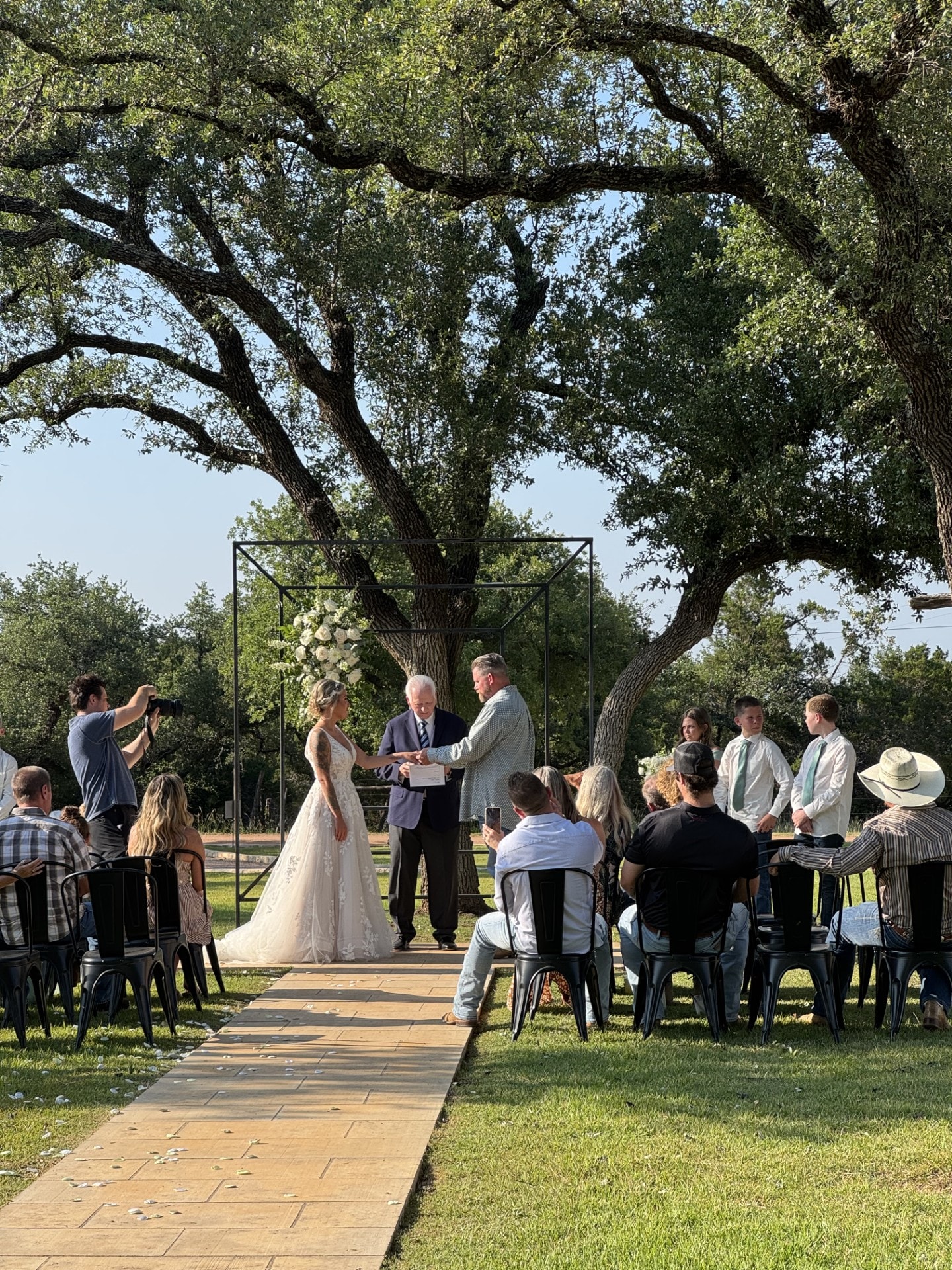 Bride and groom exchanging vows at an outdoor evening ceremony at Willow Sky Ranch — guests seated under a canopy of live oak trees on 75 private acres near Fredericksburg, Texas Hill Country