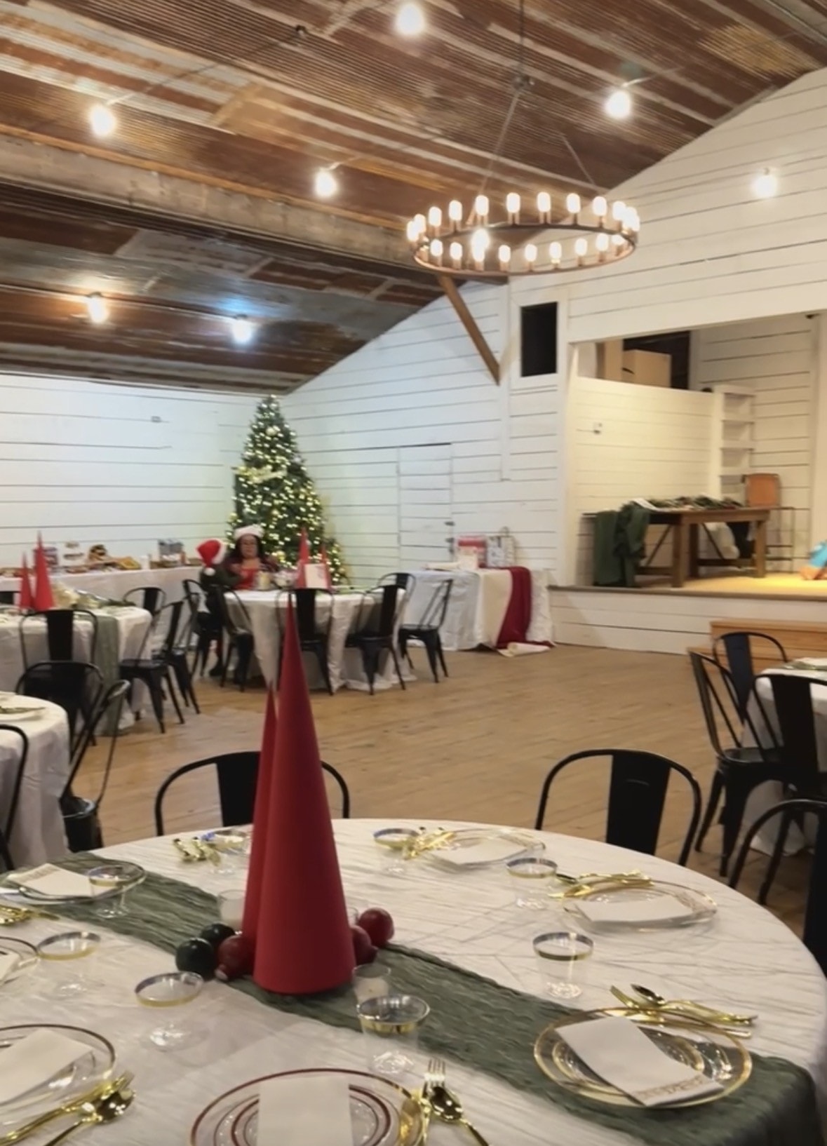 Willow Hall at Willow Sky Ranch decorated for a holiday party — round tables with white linens, red Christmas tree centerpieces, gold place settings, and a lit Christmas tree in the background near Fredericksburg, Texas Hill Country