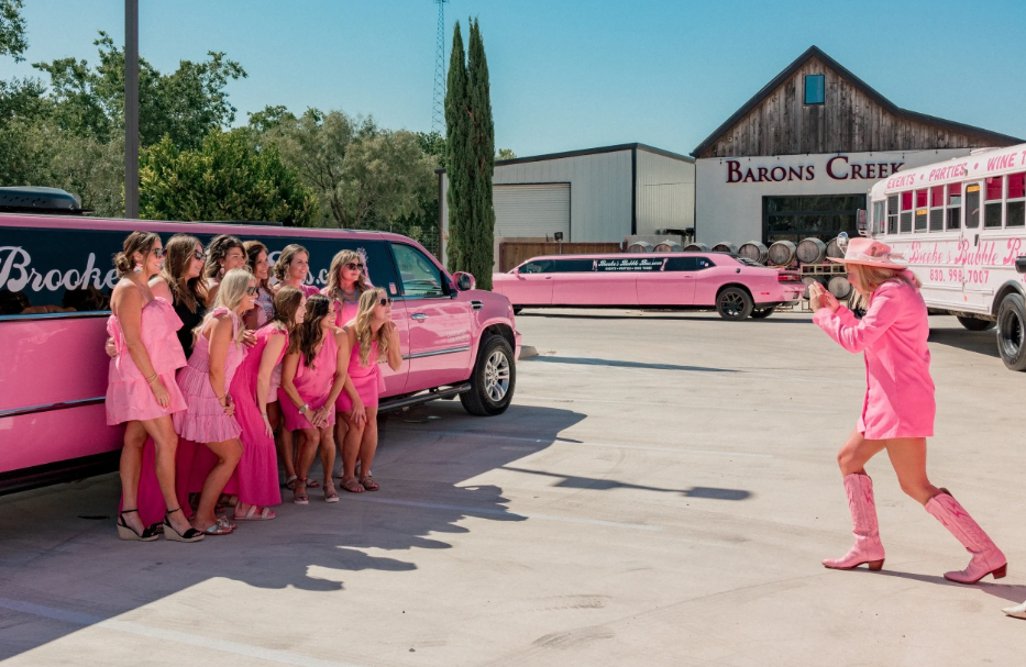 Bachelorette group in pink outfits posing with Brooke's Bubble Bus pink limo fleet at Barons Creek Vineyard in Fredericksburg, TX