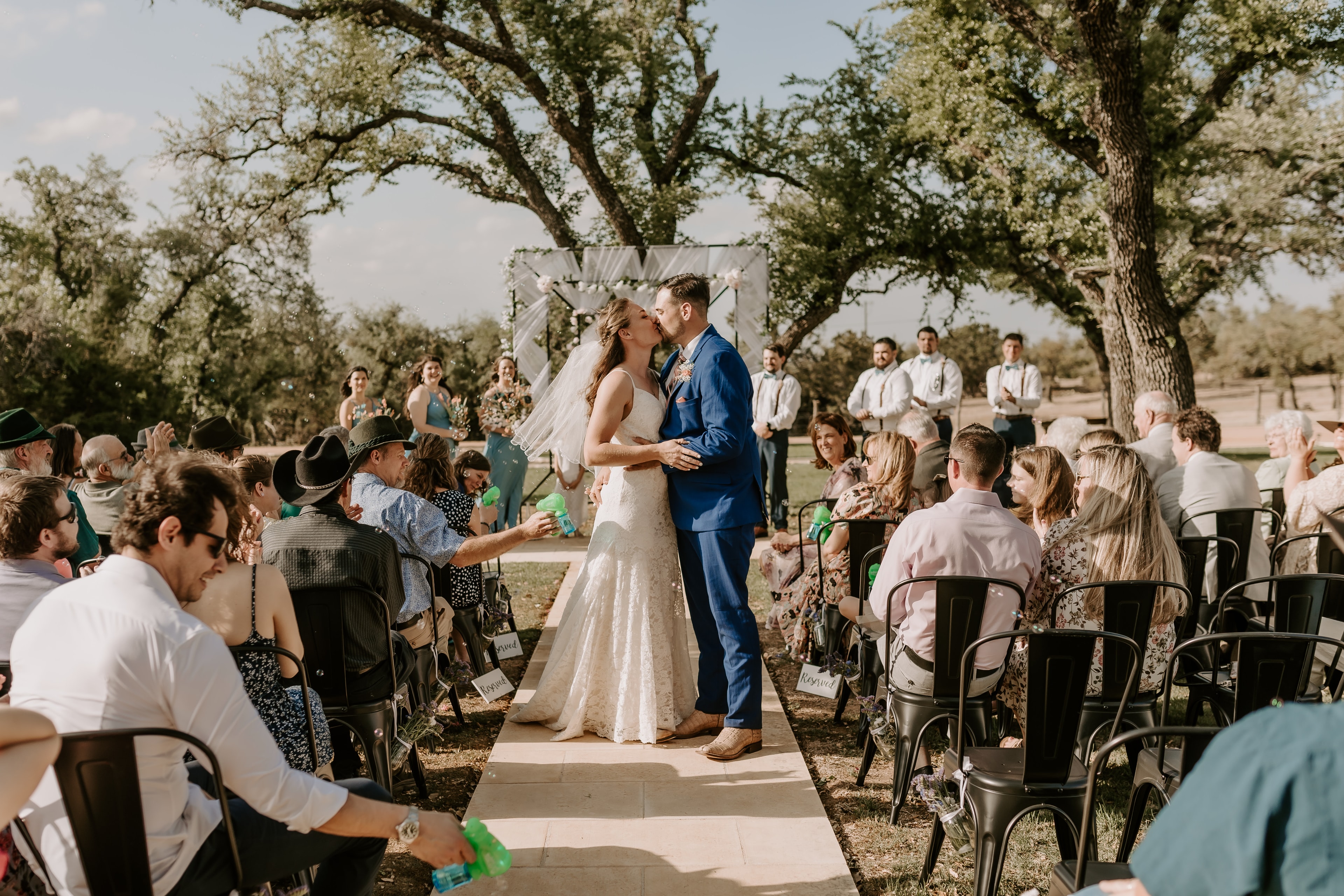 First kiss at an outdoor wedding ceremony under live oak trees at Willow Sky Ranch near Fredericksburg, TX — the kind of moment engaged couples will dream of at the First Date After the Ring experience