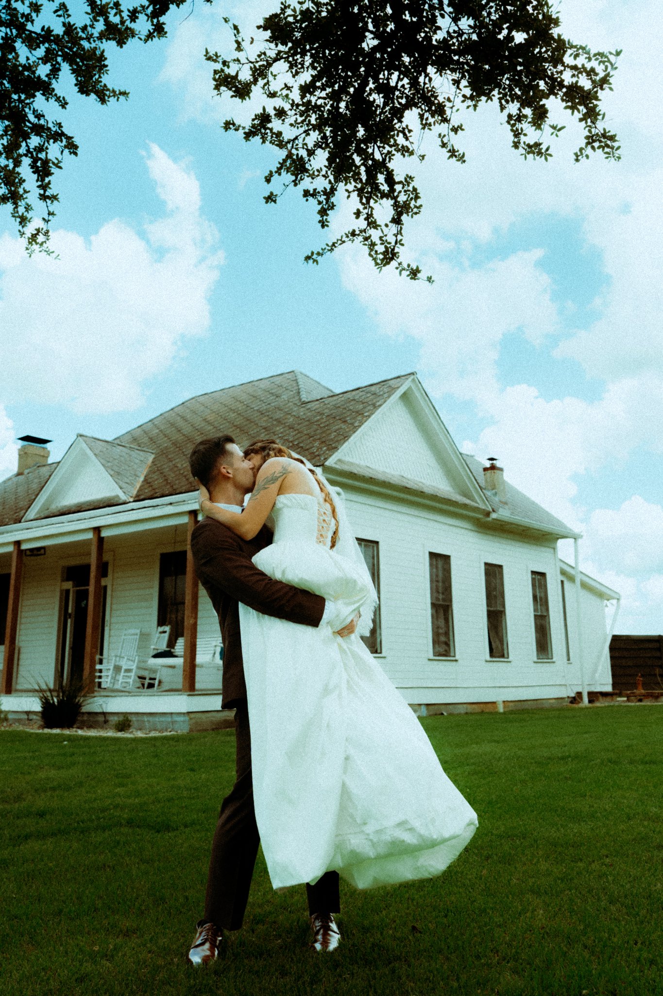 Groom lifting bride in a white off-the-shoulder gown in front of The Greathouse at Willow Sky Ranch near Fredericksburg, TX — historic 1905 white farmhouse with covered porch and live oak tree overhead, Hill Country wedding venue near Austin and San Antonio, photographed by Zach Brigham Delude Photography